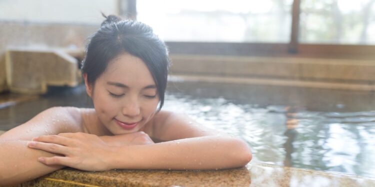 A woman rests beside a steaming indoor Tokyo onsen pool, eyes closed, surrounded by tiled walls and warm mineral water.