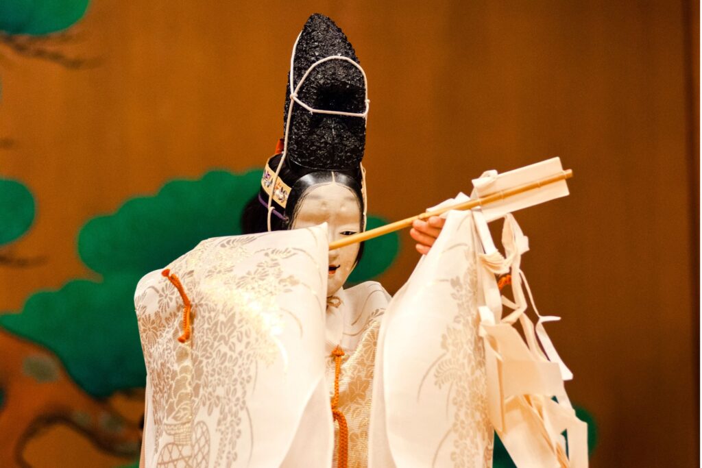 Noh theater performer in a white brocade kimono wearing a tall black mask, holding a wooden wand wrapped with white paper streamers on stage.