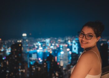 A woman holding a cocktail glass at one of Tokyo's rooftop bars, with the glowing city skyline visible behind her at dusk.