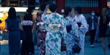 woman-yukata-night-fireworks-crowd-unsplash - TOKYOwebzine A woman in a yukata smiles while watching the Hanabi Tokyo fireworks show at night, surrounded by a crowd of festival-goers.