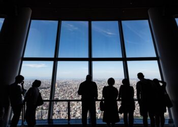 A business executive reviews documents at a Tokyo headquarters office, with a city skyline visible through the window.