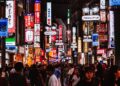 A crowded neon-lit street in Japan at night, with colorful signs and storefronts lining the sidewalk as shoppers pass by.