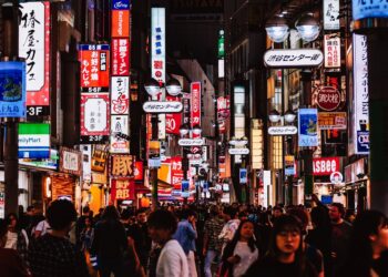 A crowded neon-lit street in Japan at night, with colorful signs and storefronts lining the sidewalk as shoppers pass by.