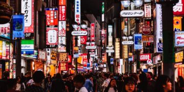 A crowded neon-lit street in Japan at night, with colorful signs and storefronts lining the sidewalk as shoppers pass by.