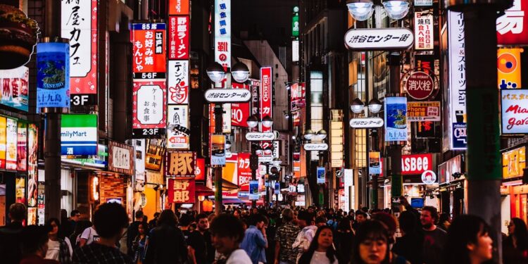 tokyo-street-fashion-unsplash - TOKYOwebzine A crowded neon-lit street in Japan at night, with colorful signs and storefronts lining the sidewalk as shoppers pass by.
