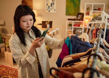 Woman with dark hair wearing a plaid shirt holds a green smartphone while selecting clothes in a warmly lit boutique.