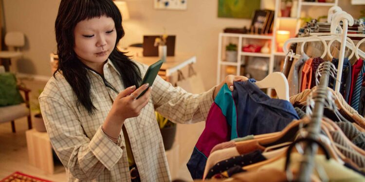 woman-browsing-vintage-clothing-rack-tokyo - TOKYOwebzine Woman with dark hair wearing a plaid shirt holds a green smartphone while selecting clothes in a warmly lit boutique.