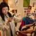 woman-browsing-vintage-clothing-rack-tokyo - TOKYOwebzine Woman with dark hair wearing a plaid shirt holds a green smartphone while selecting clothes in a warmly lit boutique.