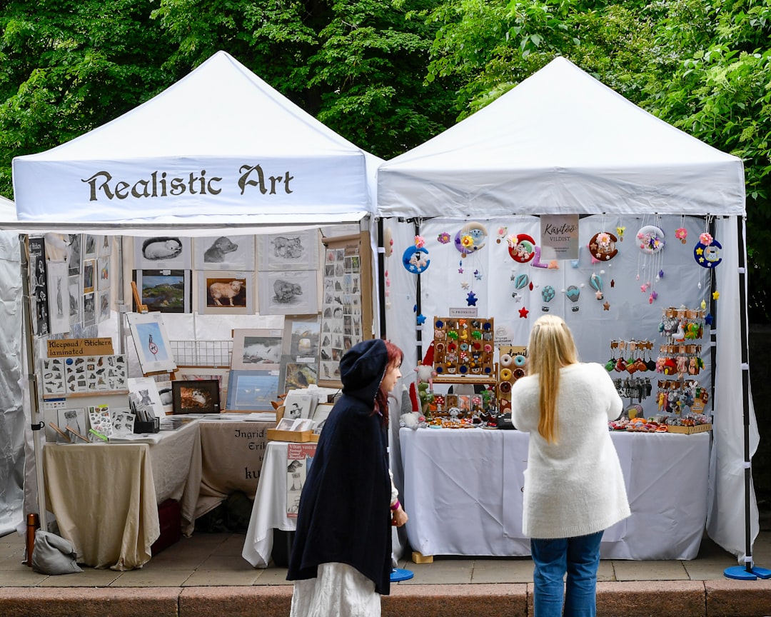Two visitors browse handmade goods and illustration work at an open-air art market in Tokyo near a waterfront venue.