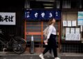 A woman dressed in loose natural Japanese fabrics walks through a shaded Tokyo street on a hot August afternoon