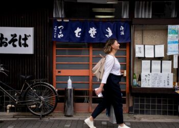 A woman dressed in loose natural Japanese fabrics walks through a shaded Tokyo street on a hot August afternoon
