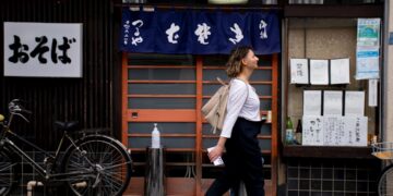 A woman dressed in loose natural Japanese fabrics walks through a shaded Tokyo street on a hot August afternoon