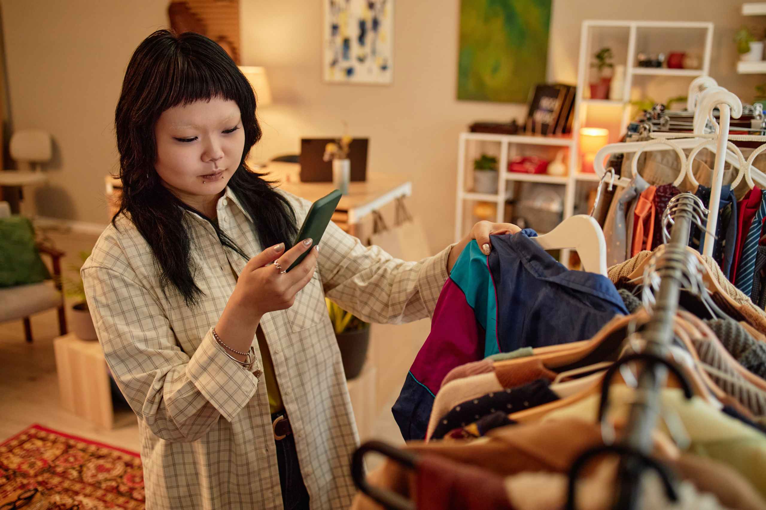 Woman with dark hair wearing a plaid shirt holds a green smartphone while selecting clothes in a warmly lit boutique.